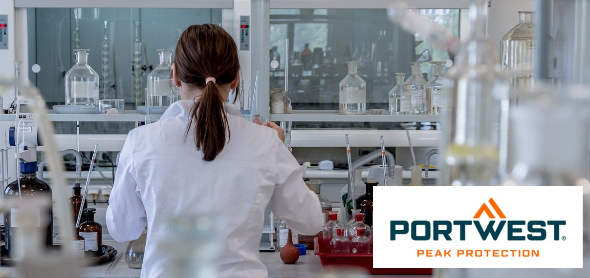 Uma mulher com cabelo castanho escuro preso para trás usa um jaleco branco e trabalha em um laboratório moderno com vários equipamentos de laboratório e frascos de produtos químicos. No canto inferior direito da imagem está o logotipo “Portwest Peak Protection”.
