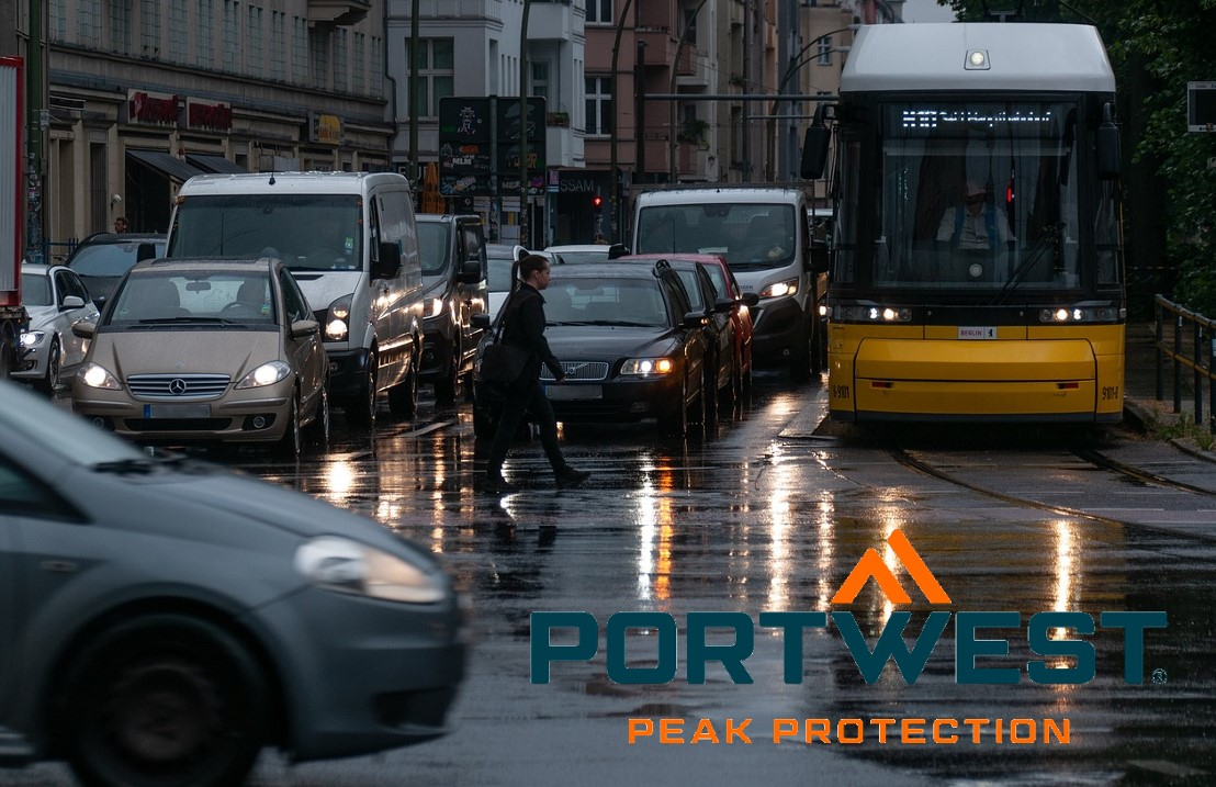 Cena de rua à chuva. Vários carros e autocarros estão estacionados num cruzamento com os faróis acesos. O logótipo da empresa Portwest em laranja e azul encontra-se no canto inferior direito da imagem. Existe um link que leva às nossas capas de chuva.