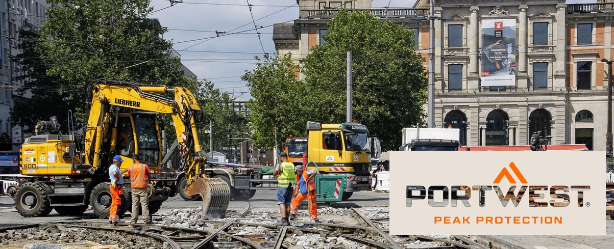 Trabalhadores da construção civil com roupas de segurança cor de laranja trabalham num estaleiro de obras na cidade. Escavadoras, camiões e um edifício histórico podem ser vistos ao fundo. O logótipo da Portwest e o slogan “Peak Protection” são apresentados na parte inferior da imagem.