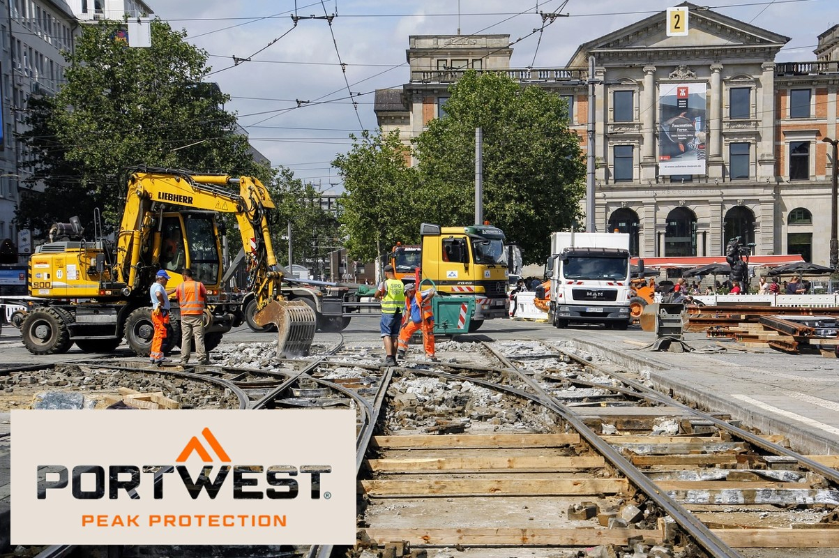 Trabalhadores da construção civil com roupas de segurança cor de laranja trabalham num estaleiro de obras na cidade. Escavadoras, camiões e um edifício histórico podem ser vistos ao fundo. O logótipo da Portwest e o slogan “Peak Protection” são apresentados na parte inferior da imagem. Há um link que leva à página do artigo da tampa de refrigeração.